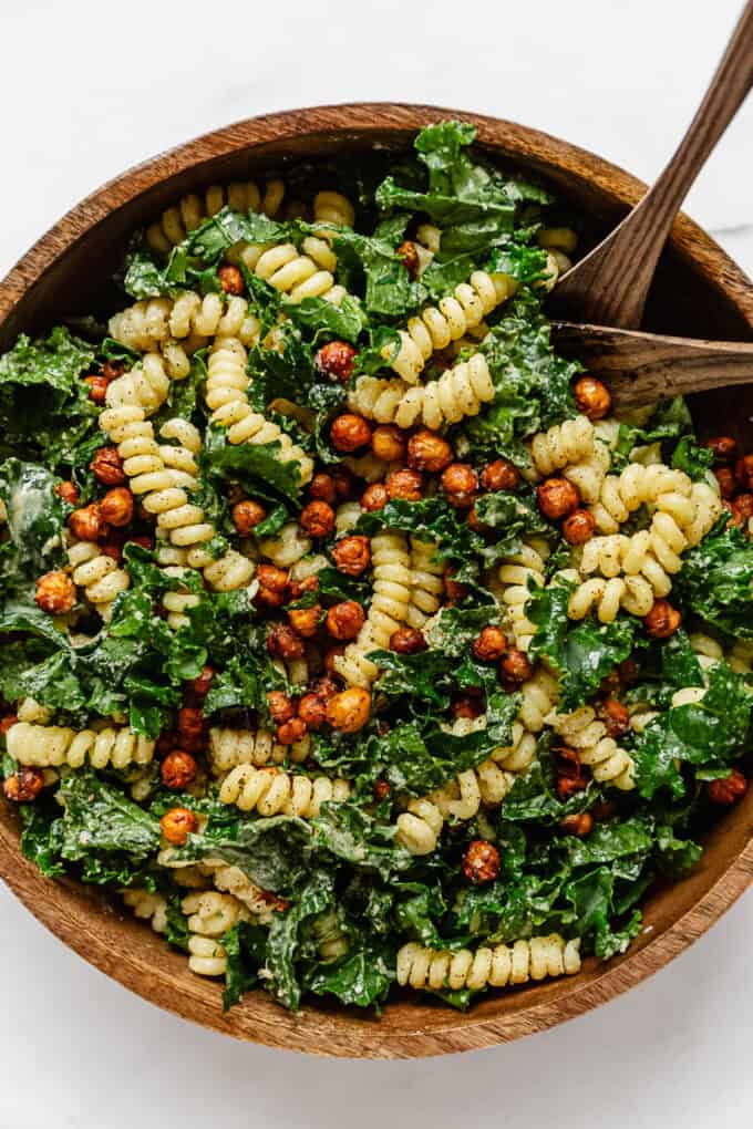 kale caesar pasta salad in a wood mixing bowl with 2 wood spoons