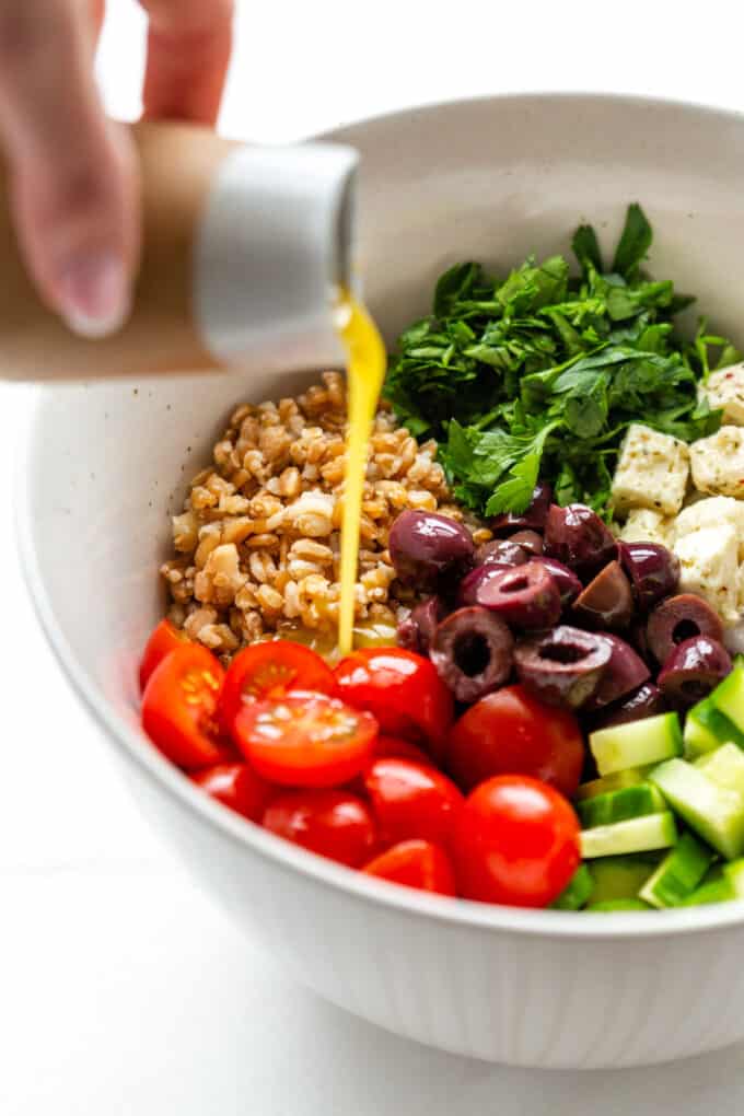 Greek salad dressing being poured into a bowl of a farro salad