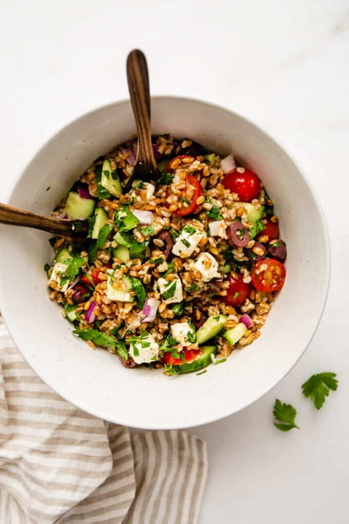 Greek farro salad in a bowl with serving spoons and a striped napkin on the side